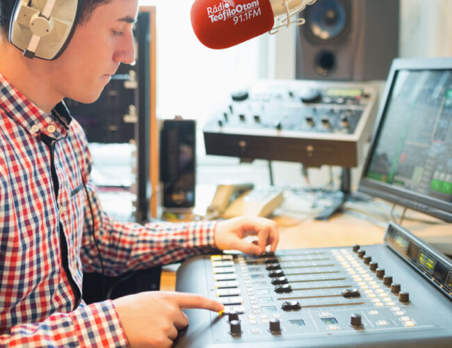 Radio host wearing headphones operating sound mixer on table in studio