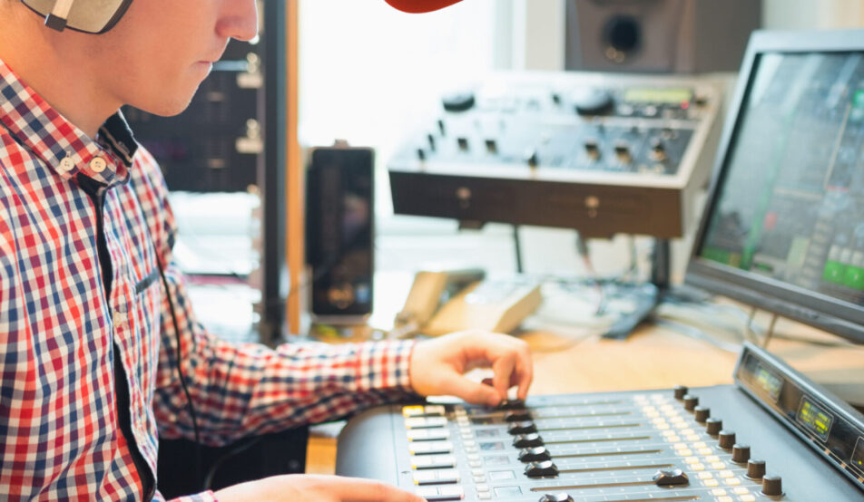 Radio host wearing headphones operating sound mixer on table in studio